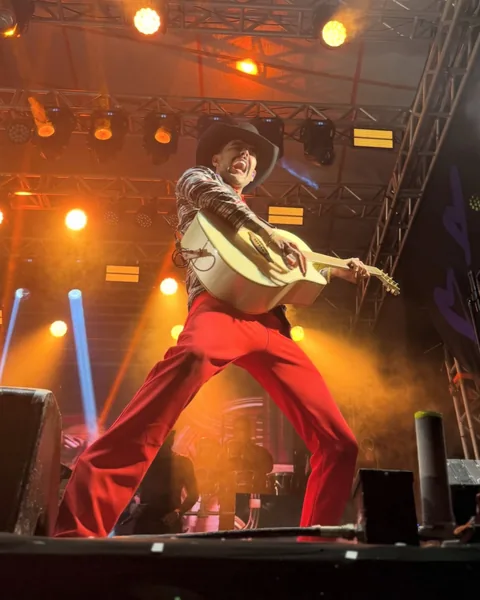 Cantor de chapéu e calça vermelha tocando violão no palco, com treliça de luz e refletores alaranjados
